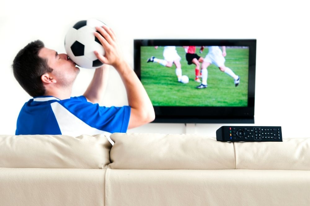 A man kissing the soccer ball is sitting in front of the TV 