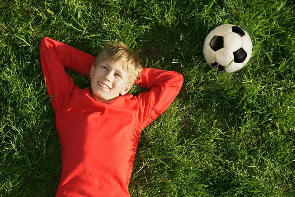 a soccer player wearing red jersey lying on grass with a soccer ball next to him-min