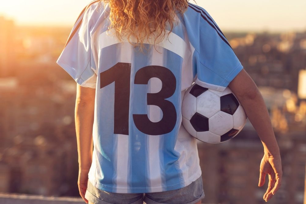 girl wearing soccer jersey number 13 with shorts, holding a soccer ball in her ampit