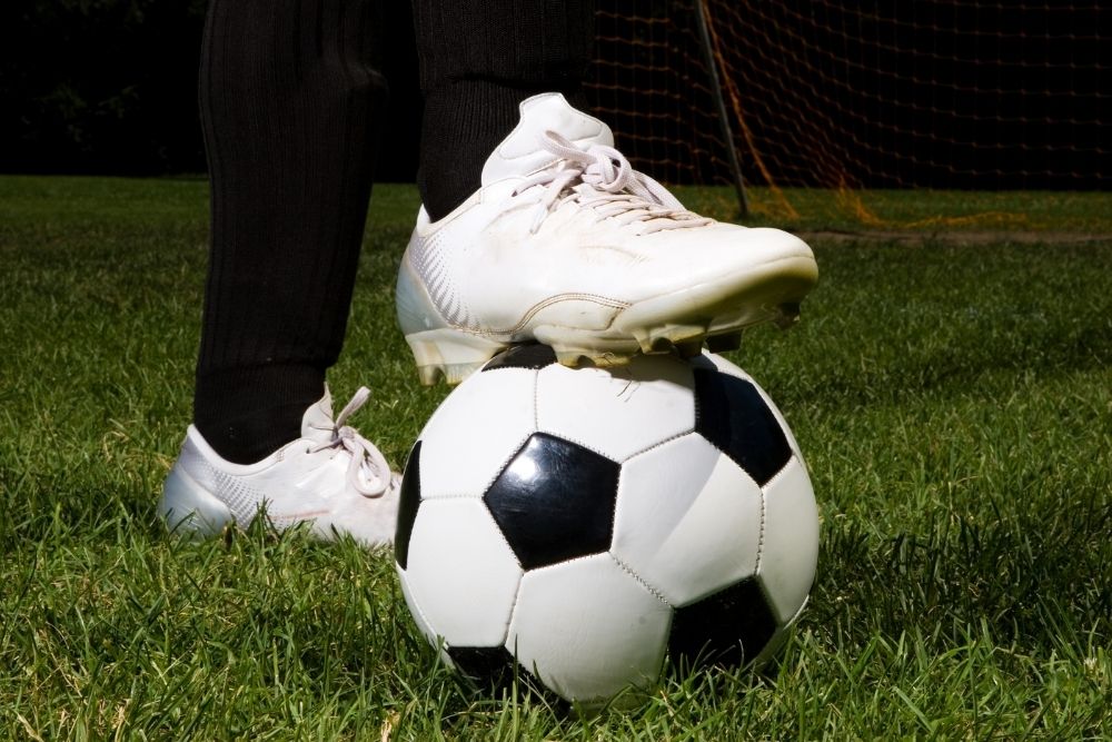 a person wearing white soccer cleats on grass, put a foot on soccer ball