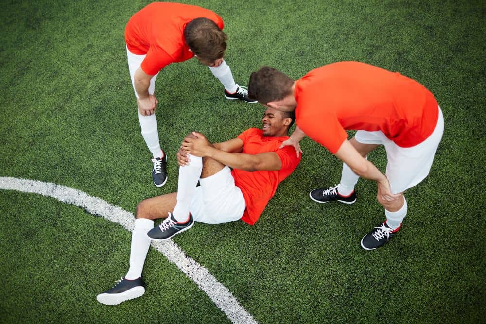 soccer player is in pain and holding his knee with his teammates gather around